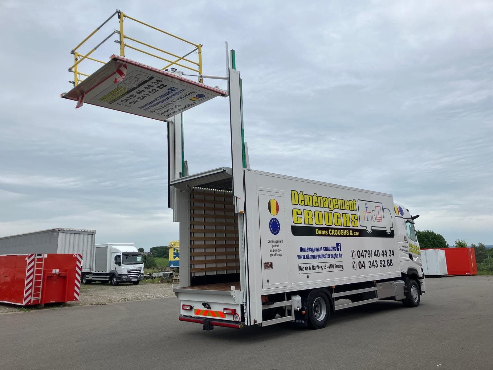 Camion Croughs vu de l'arrière, caisse ouverte et plateforme du lift intégré levée au-dessus du toit, sur un parking de dépôt