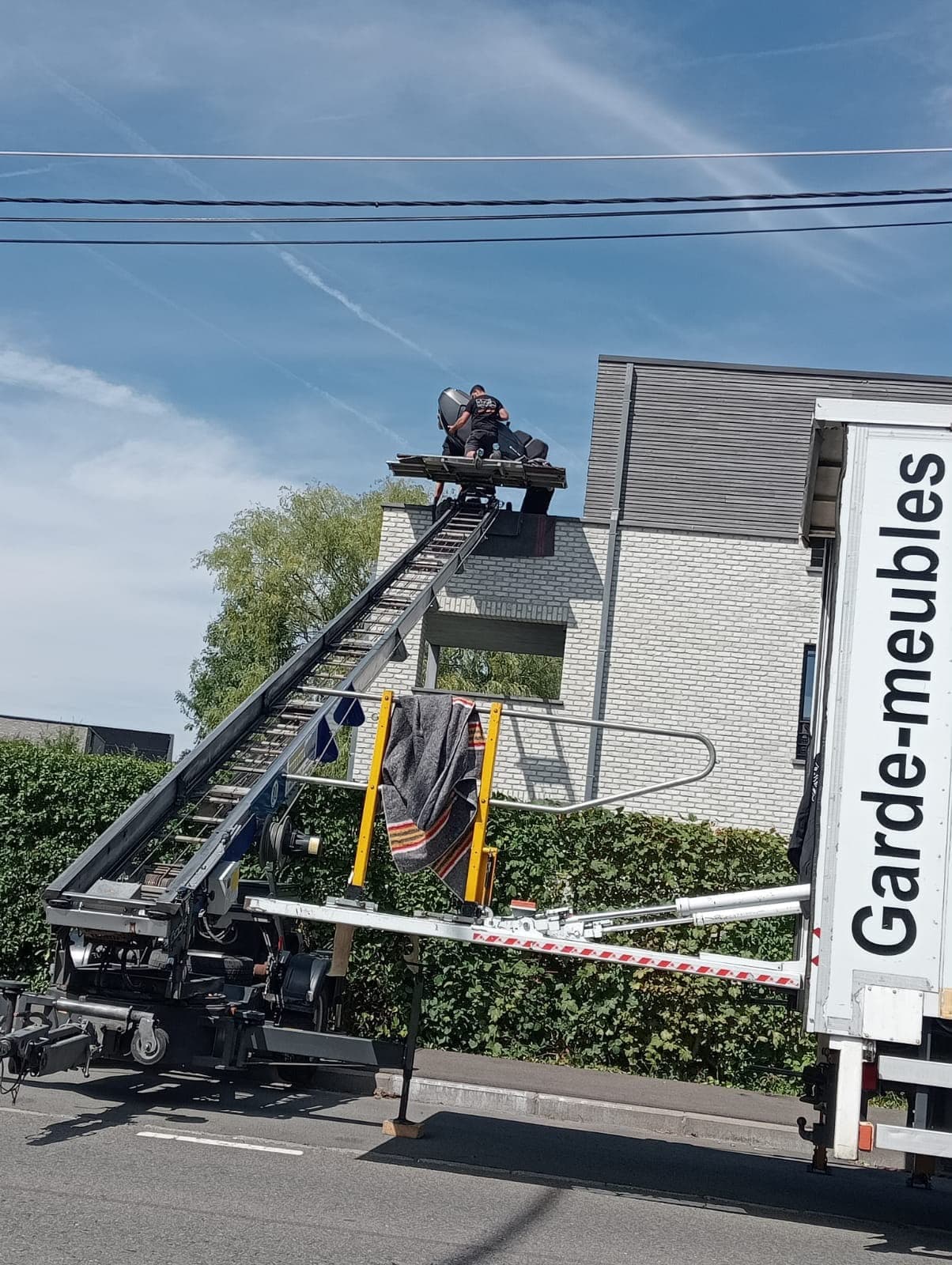 Déménageur Croughs, de dos, sur la plateforme du lift en haut de l'échelle, guidant un fauteuil vers l'étage d'une maison