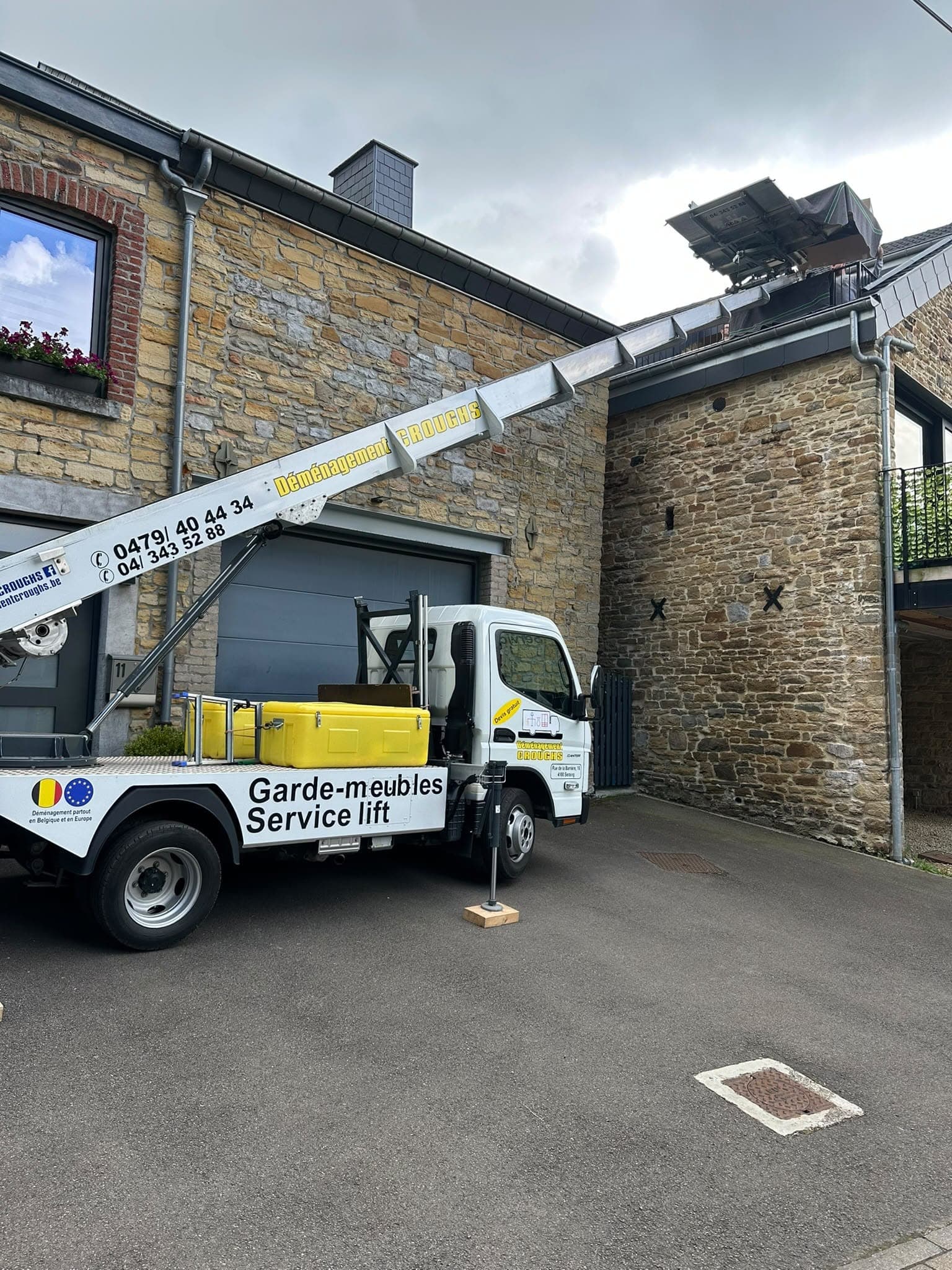 Camion « Garde-meubles, Service lift » de Croughs devant une maison en pierre, échelle du lift posée jusqu'au toit