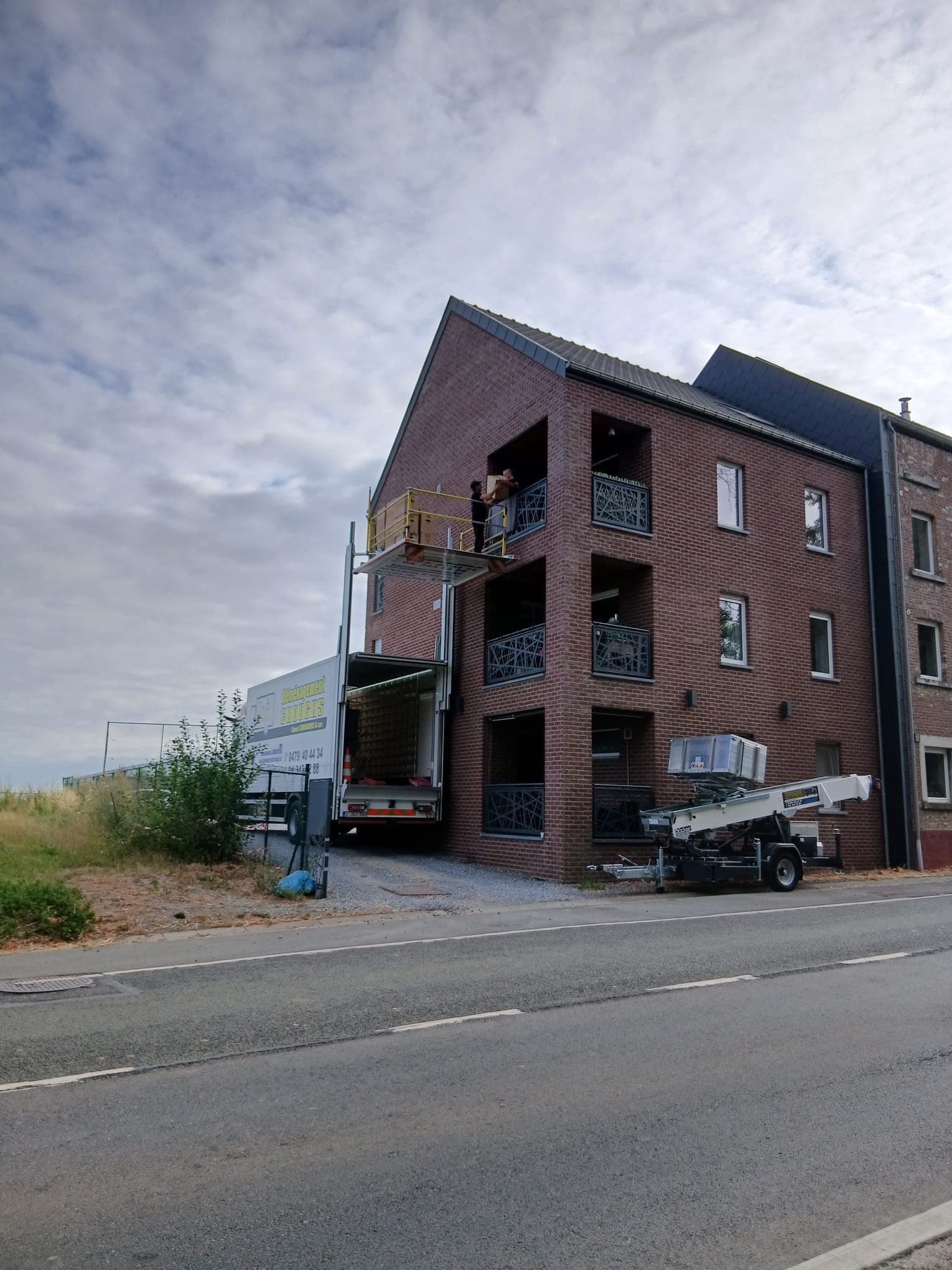 Lift Croughs déployé contre un immeuble en briques : deux déménageurs déchargent des cartons sur la plateforme à hauteur d'un balcon, camion ouvert en dessous