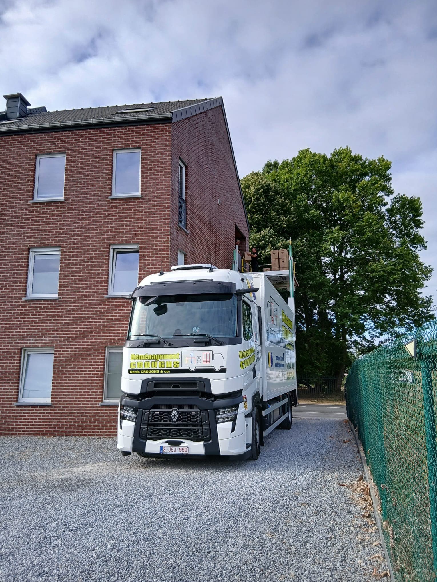 Camion Croughs Déménagement garé devant un immeuble en briques, plateforme du lift levée à l'étage avec des cartons et deux déménageurs