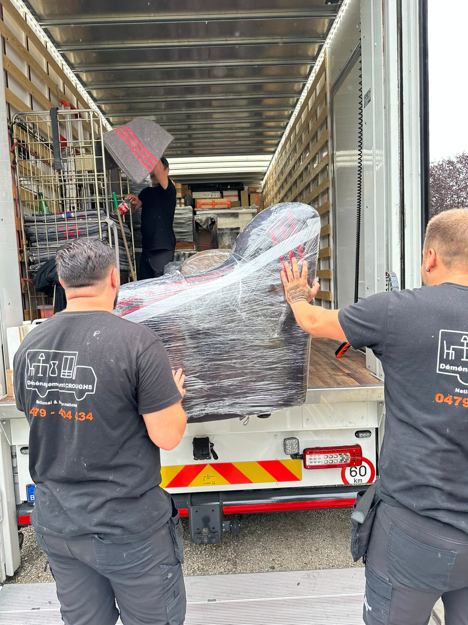 Deux déménageurs Croughs, de dos, poussent un fauteuil emballé sous film dans un camion déjà chargé de meubles protégés