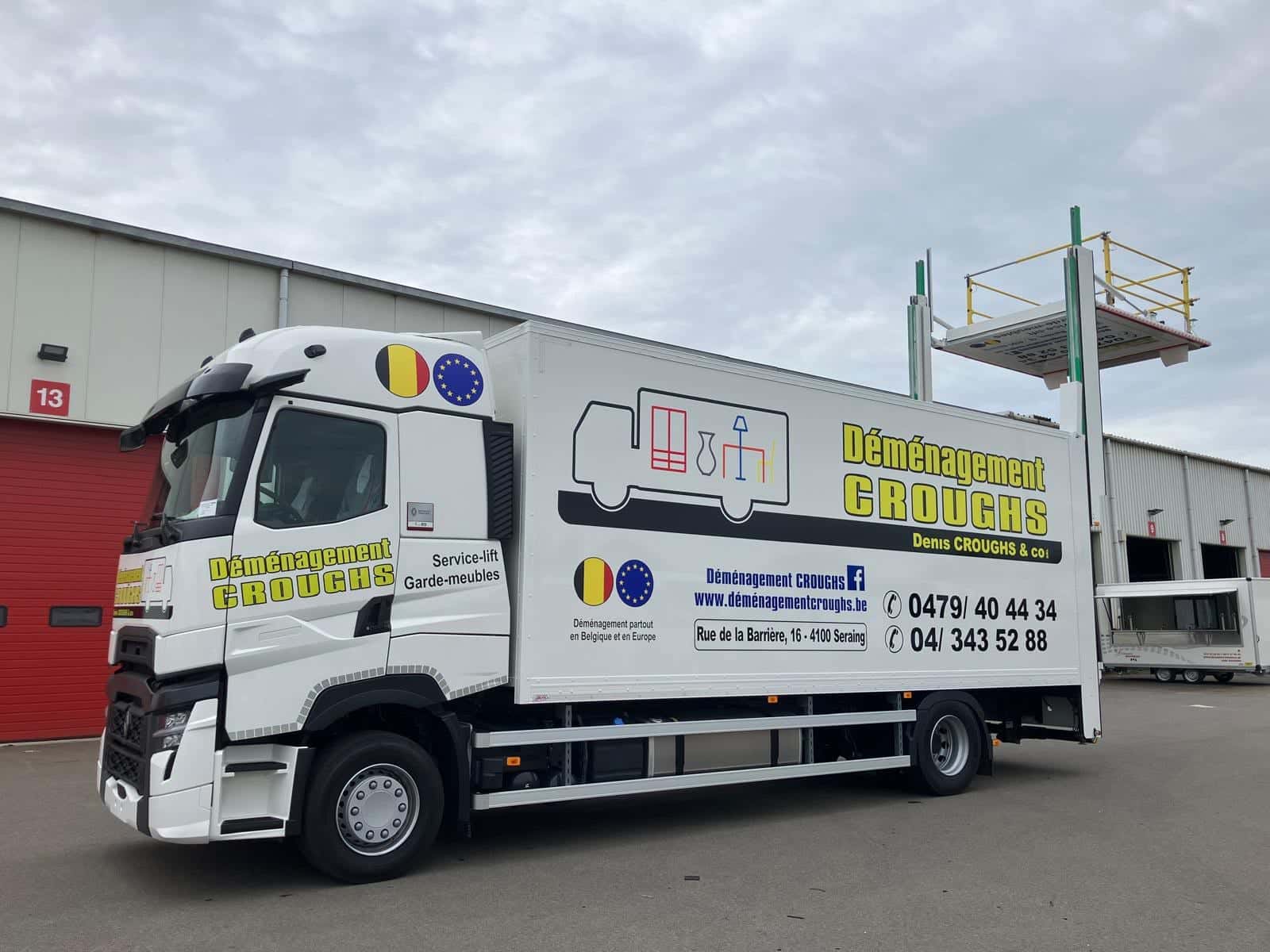 Camion Déménagement Croughs de profil devant un dépôt, plateforme du lift intégré levée au-dessus de la caisse